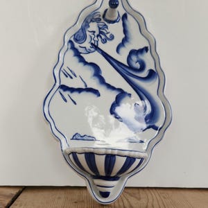 May include: A white ceramic wall-mounted holy water font featuring a blue hand-painted design of a face blowing wind from the clouds. The piece has a scalloped edge, a small basin at the bottom with blue vertical stripes, and a top hanging hole.