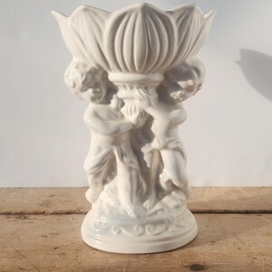 Peut inclure: Un vase en céramique blanche orné d'une fleur de lotus soutenue par trois chérubins. Le vase présente une surface texturée et détaillée et repose sur une surface en bois. Le design est classique et décoratif.
