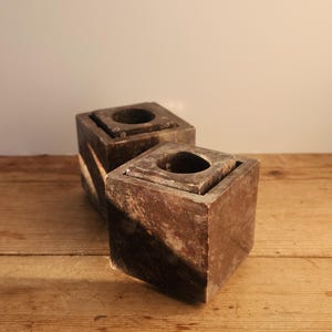May include: Two square, brown marble candle holders with a central circular opening. The candle holders are placed on a wooden surface. The marble has a natural, veined pattern.