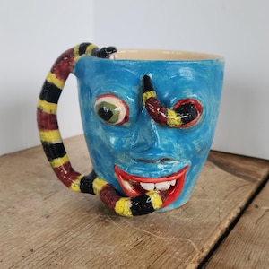 May include: A blue ceramic mug featuring a sculpted face with a wide red mouth, white teeth, and a single visible green eye. A snake with red, yellow, and black bands forms the handle, wrapping around the side and resting inside the mouth.