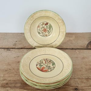 Puede incluir: Platos color crema con un diseño floral central, bordes verdes y anillos negros. Una pila de platos y un plato separado se muestran sobre una superficie de madera. Los platos tienen aproximadamente 20 cm de diámetro.