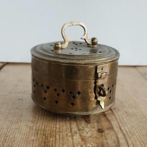 May include: A round brass container with a hinged lid and a small handle on top. The sides and lid feature small, perforated cutouts in a cross-like pattern. A metal latch mechanism is attached to the side to secure the lid to the base.