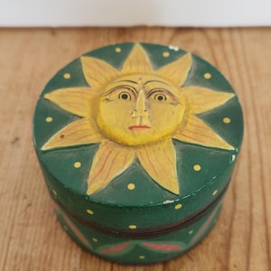 Könnte beinhalten: Eine runde, handbemalte Holzbox mit einem Sonnendesign auf dem Deckel. Die Sonne hat ein gelbes Gesicht und Strahlen, vor einem grünen Hintergrund mit gelben Punkten. Die Box steht auf einer Holzoberfläche.