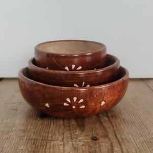 May include: A set of three wooden bowls in varying sizes, stacked on top of each other. The bowls are a rich brown color with a light-colored floral design. The bowls are on a wooden surface.