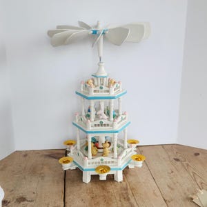 Puede incluir: Un carrusel blanco y azul de varios niveles con figuras, coronado por un abanico. El carrusel tiene un borde azul claro y candelabros dorados. El carrusel está sobre una mesa de madera.