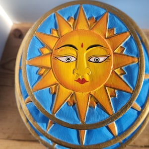 Könnte beinhalten: Holzschnitzerei eines Sonnengesichts mit gelbem Gesicht, orangefarbenen Strahlen und blauem Hintergrund. Die Sonne hat detaillierte Augen, einen roten Punkt auf der Stirn und einen roten Mund. Die Sonne ist von einem goldenen Rand umgeben.