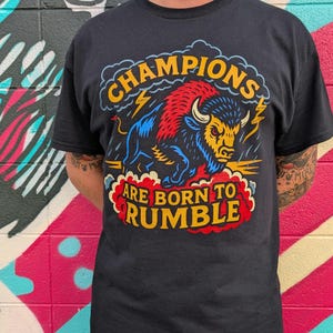 Op de afbeelding: Zwart t-shirt met een kleurrijke afbeelding van een bizon en de tekst "CHAMPIONS ARE BORN TO RUMBLE". De bizon is afgebeeld in blauw, rood en geel, met bliksemflitsen en wolken. De tekst is in geel en rood.