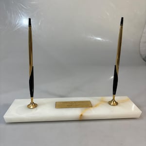 Könnte beinhalten: Ein Stifthalter mit zwei goldfarbenen Stiften mit schwarzen Akzenten, auf einem weißen Marmorsockel. Ein kleines goldenes Schild mit graviertem Text befindet sich in der Mitte des Sockels. Die Stifte haben ein schlankes, modernes Design.
