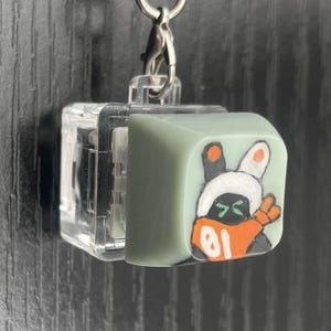 May include: A keychain featuring a small, light green cube with a cartoon character design. The character has a white bunny-like head with orange accents and the number "01". The cube is attached to a silver chain.