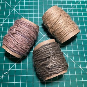 May include: Three spools of twine in varying shades of brown and gray are arranged on a green cutting mat. The twine is tightly wound around cardboard tubes, showcasing a rustic aesthetic suitable for crafts and DIY projects.