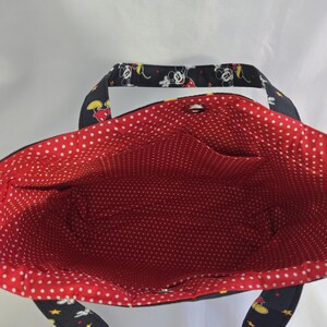 May include: A black tote bag with a red polka dot interior. The exterior features a pattern of Mickey Mouse, stars, and yellow accents. The bag has two pockets inside and a snap closure.
