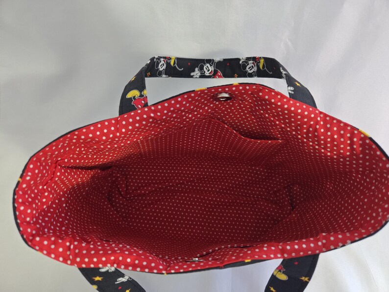 May include: A black tote bag with a red polka dot interior. The exterior features a pattern of Mickey Mouse, stars, and yellow accents. The bag has two pockets inside and a snap closure.