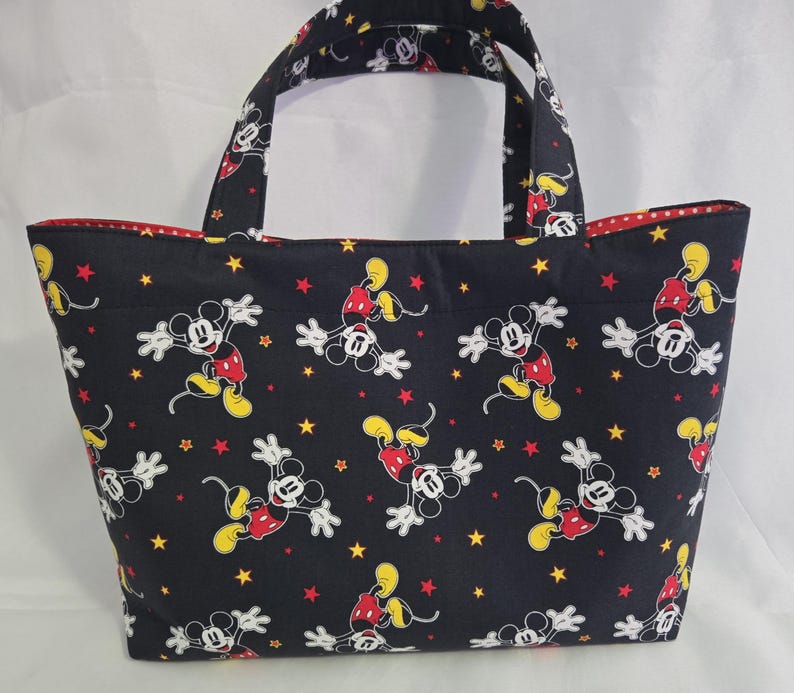 May include: A black tote bag featuring a repeating pattern of Mickey Mouse in various poses, along with yellow and red stars. The bag has red polka dot lining and matching handles.