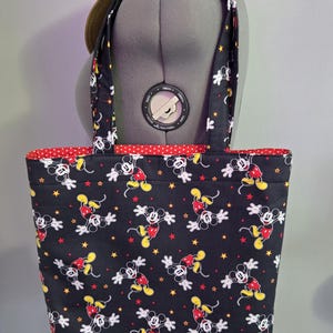 May include: Black tote bag featuring a repeating pattern of Mickey Mouse in red, yellow, and white on a black background. The bag has red polka dot lining and matching handles. A straw hat with a black band rests on top.