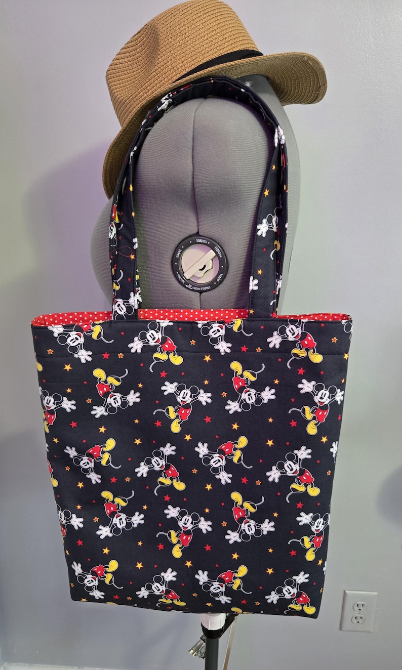 May include: Black tote bag featuring a repeating pattern of Mickey Mouse in red, yellow, and white on a black background. The bag has red polka dot lining and matching handles. A straw hat with a black band rests on top.