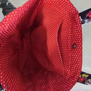 May include: A black tote bag with a red polka dot interior. The bag has a black exterior with cartoon character designs and a small interior pocket. The interior is red with white polka dots.