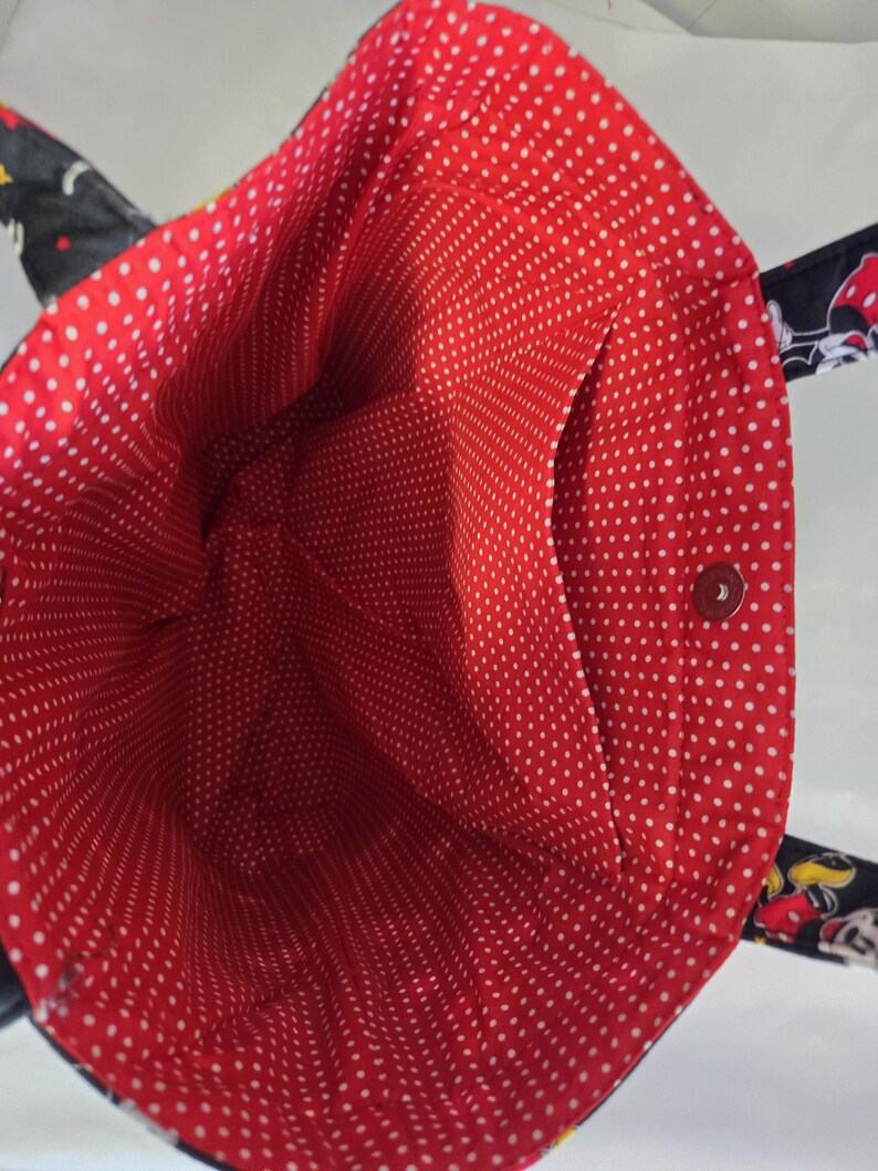 May include: A black tote bag with a red polka dot interior. The bag has a black exterior with cartoon character designs and a small interior pocket. The interior is red with white polka dots.