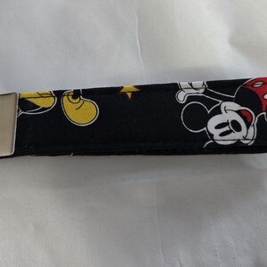 May include: Black fabric key fob featuring a Mickey Mouse pattern. The design includes Mickey Mouse in various poses with yellow shoes, red shorts, and white gloves. A silver metal key ring and clasp are attached.