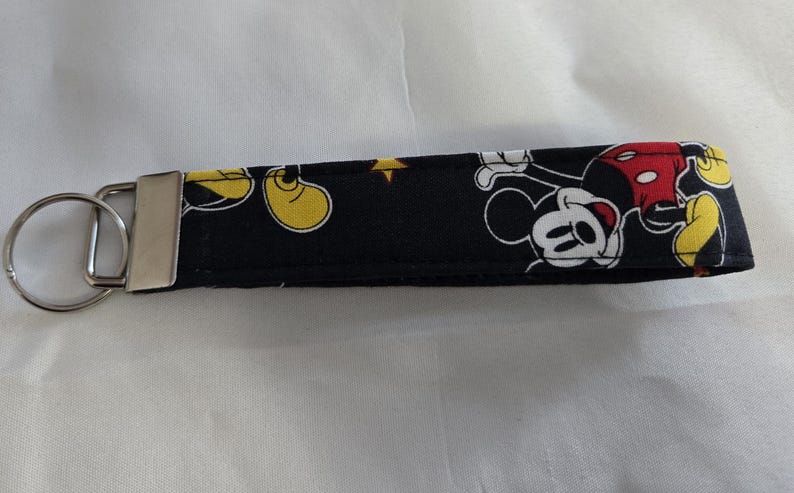 May include: Black fabric key fob featuring a Mickey Mouse pattern. The design includes Mickey Mouse in various poses with yellow shoes, red shorts, and white gloves. A silver metal key ring and clasp are attached.