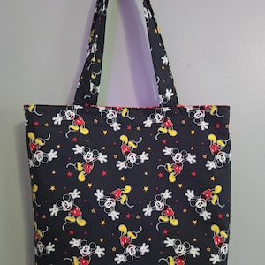 May include: Black tote bag featuring a repeating pattern of Mickey Mouse in various poses, with red, yellow, and white accents, and yellow and red stars. The bag has black handles and a red interior lining.