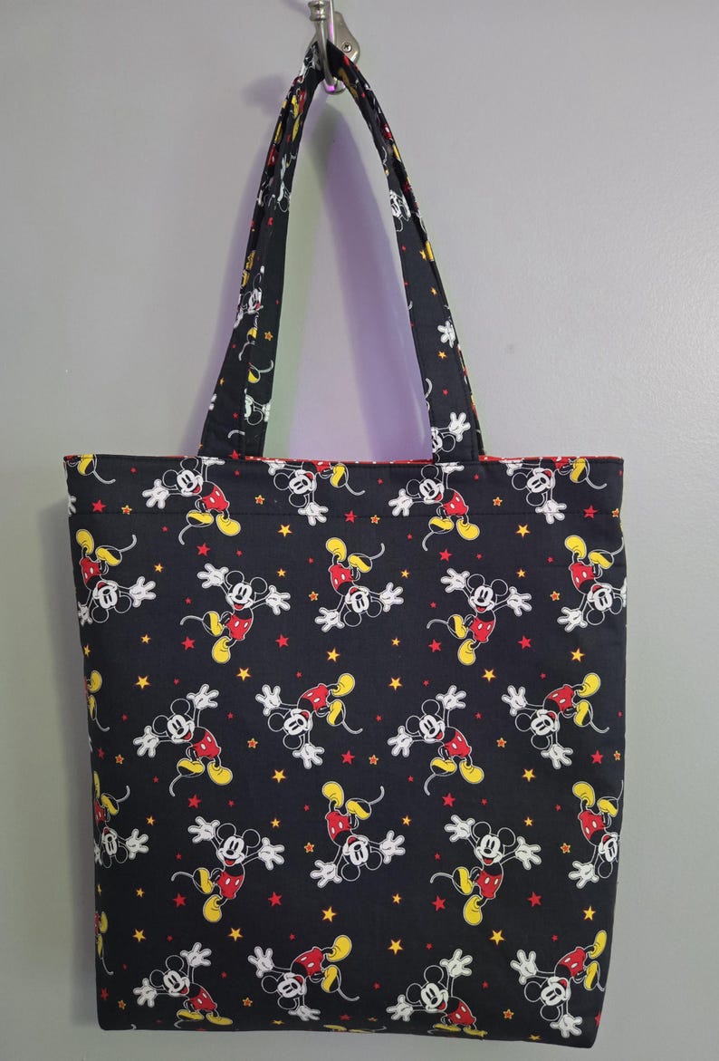 May include: Black tote bag featuring a repeating pattern of Mickey Mouse in various poses, with red, yellow, and white accents, and yellow and red stars. The bag has black handles and a red interior lining.