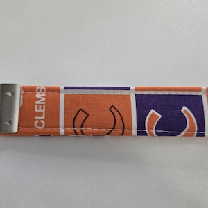 May include: A fabric keychain featuring orange and purple squares with the word "CLEMS" and a stylized "C" logo. The keychain has a silver metal clasp and ring.