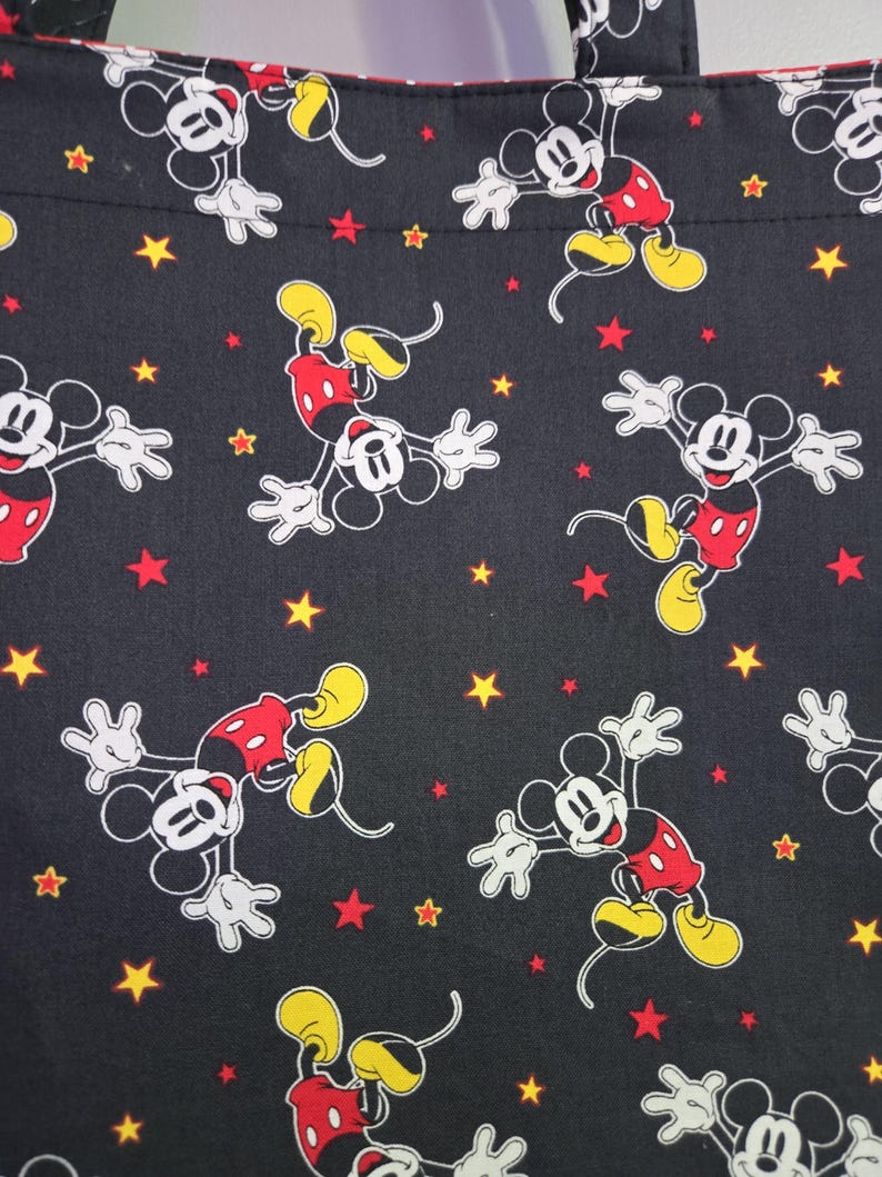 May include: Black tote bag featuring a repeating pattern of Mickey Mouse in various poses. The design includes red, yellow, and white Mickey Mouse figures, along with red and yellow stars scattered across the black background.