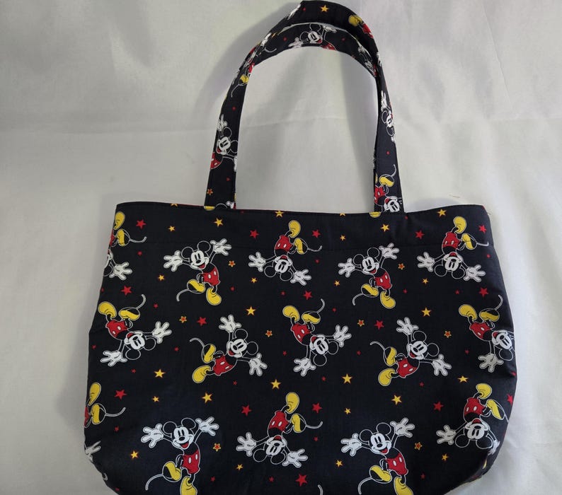 May include: Black tote bag featuring a repeating pattern of Mickey Mouse in various poses with yellow and red stars. The bag has two handles and is made of fabric.