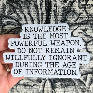 May include: A white sticker with black text that reads: "KNOWLEDGE IS THE MOST POWERFUL WEAPON. DO NOT REMAIN WILLFULLY IGNORANT DURING THE AGE OF INFORMATION." The sticker is held by a hand.
