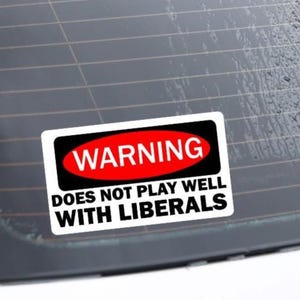 May include: A white warning sticker with red and black text on a car window. The sticker reads "Warning Does Not Play Well With Liberals".