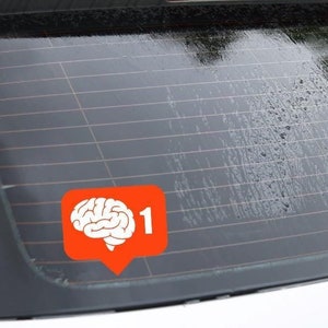 Peut inclure: Une voiture blanche avec un autocollant sur la vitre arrière. L'autocollant est orange et représente une icône de cerveau blanche avec le chiffre "1" à côté.