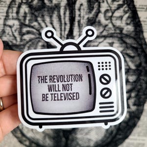 May include: A black and white sticker in the shape of a retro television. The screen displays the text "THE REVOLUTION WILL NOT BE TELEVISED". The sticker has a white border and antenna.