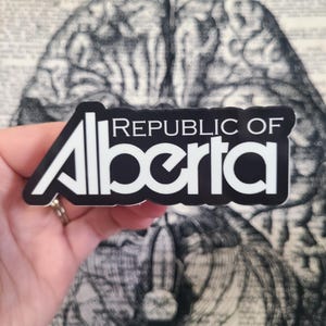 May include: A black and white sticker with the text "REPUBLIC OF Alberta" in a bold, sans-serif font. The sticker has a white border and is held in front of a vintage brain illustration.