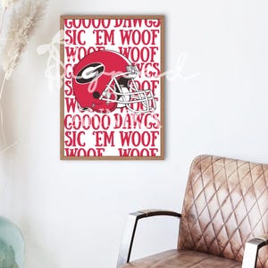 May include: A framed print with a red American football helmet and the text "GOOOD DAWGS SIC 'EM WOOF WOOF" in red. The print is in a brown frame and hangs on a white wall. The helmet has a white facemask.