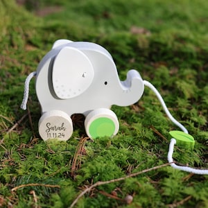 Könnte beinhalten: Ein graues, hölzernes Elefanten-Ziehspielzeug mit grünen Rädern und einem weißen Seil. Der Elefant hat eine personalisierte Inschrift auf dem unteren Rad, die "Sarah 11.11.24" lautet.