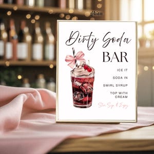 May include: A framed sign with the words "Dirty Soda BAR" in elegant script. The sign features an illustration of a soda drink with ice cubes, whipped cream, and cherries. Instructions on how to make the drink are listed below the illustration.
