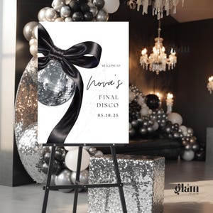 Peut inclure: Un chevalet noir présente une affiche blanche avec l'inscription « Welcome to Nova's Final Disco 05.18.25 ». L'affiche présente une boule à facettes et un ruban noir. L'arrière-plan comprend des ballons et un cube à paillettes.