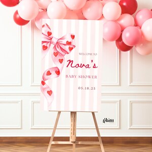 Puede incluir: Un letrero de baby shower sobre un caballete, con un fondo a rayas rosas y blancas. El letrero tiene un lazo de acuarela con corazones rojos y el texto "Welcome to Nova's Baby Shower 05.18.25". Globos rosas y rojos en el fondo.