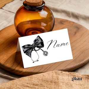 Puede incluir: Tarjeta de lugar blanca con un lazo negro y una ilustración de estetoscopio. El texto "Nombre" está impreso en la tarjeta.