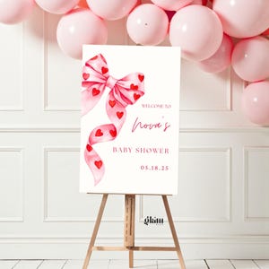 May include: A white baby shower welcome sign featuring a watercolour bow design in pink and red, decorated with heart motifs. The sign reads "Welcome to Nora's Baby Shower" with the date 05.18.25.