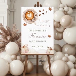 May include: A baby shower sign on a wooden easel. The sign features a coffee cup with a heart design, fall leaves, and the text "A baby is brewing." It also includes the name "Nova's" and the date 04.15.25.
