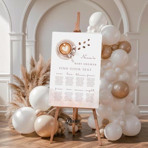 May include: A baby shower seating chart on an easel, featuring a coffee cup design with a heart-shaped latte art. The sign reads "Nova's Baby Shower" and "Find Your Seat." Balloons in white and gold hues decorate the scene, with pampas grass and a wooden floor.