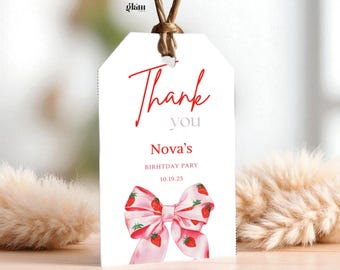 May include: A white gift tag with the words "Thank you" in red script. Below, it reads "Nova's BIRTHDAY PARTY 10.19.25". A pink bow with strawberry illustrations adorns the bottom. The tag is suspended by a brown string.