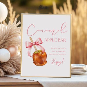 May include: A framed sign with the words "Caramel Apple Bar" in pink script. An illustration of a caramel apple with a pink bow is featured. The sign includes instructions: "Select an apple, dip in caramel, add your topping, enjoy!"