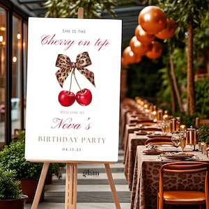 May include: A white sign on an easel with the text "She is the Cherry on top" and "Welcome to Nova's Birthday Party 04.15.25". The sign features a watercolor illustration of two red cherries and a leopard print bow.