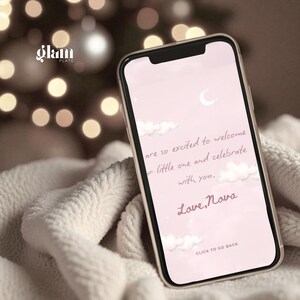 May include: A smartphone displays a pink screen with the text "are so excited to welcome our little one and celebrate with you. Love, Nova." The phone is in a gold case, resting on a white, textured blanket. The background features blurred lights.