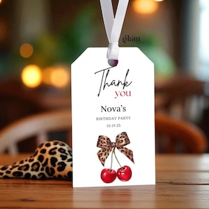May include: A white gift tag with a white ribbon features the words "Thank you" in black script and red text. Below, it reads "Nova's Birthday Party 10.19.25." A leopard print bow and two red cherries complete the design.
