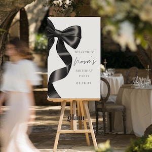 May include: A sign on a wooden easel with the text "Welcome to Nova's Birthday Party 05.18.25". The sign features a large black bow and ribbon design against a light background. The sign is displayed outdoors.