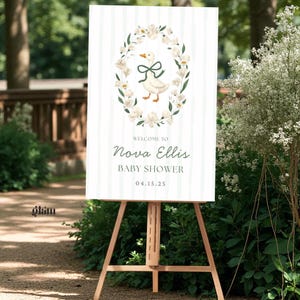 May include: A baby shower welcome sign on a wooden easel. The sign features a white background with vertical stripes and a floral wreath surrounding a cartoon goose. The text reads "Welcome to Nova Ellis Baby Shower 04.15.25".