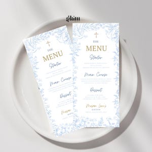 May include: Two baptism menu cards with a light blue floral design and gold text, displayed on a white plate. The cards feature the text "THE MENU", "Starter", "Main Course", and "Dessert". The menu is for "Megan Levis Baptism".